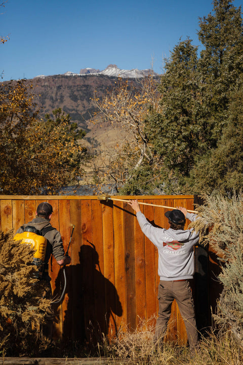 Two people brushing and spraying a wooden fence in a mountain landscape.